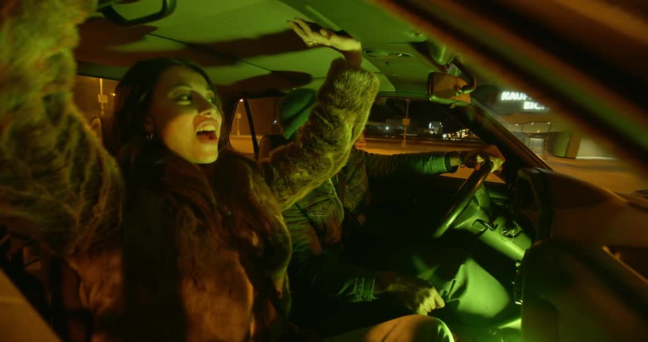 A man and woman drive around town and the woman is laughing while putting her window down