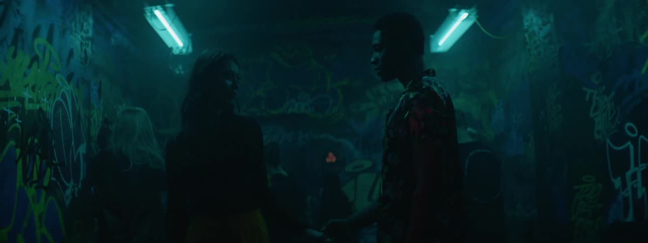 A young male and female couple stand and face each other within a dance club lit by strobing lights