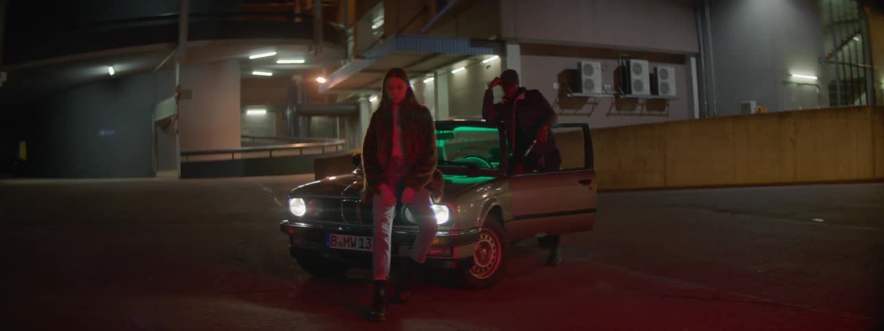 A girl leans on the hood of a car as a man stands at the door and looks at her