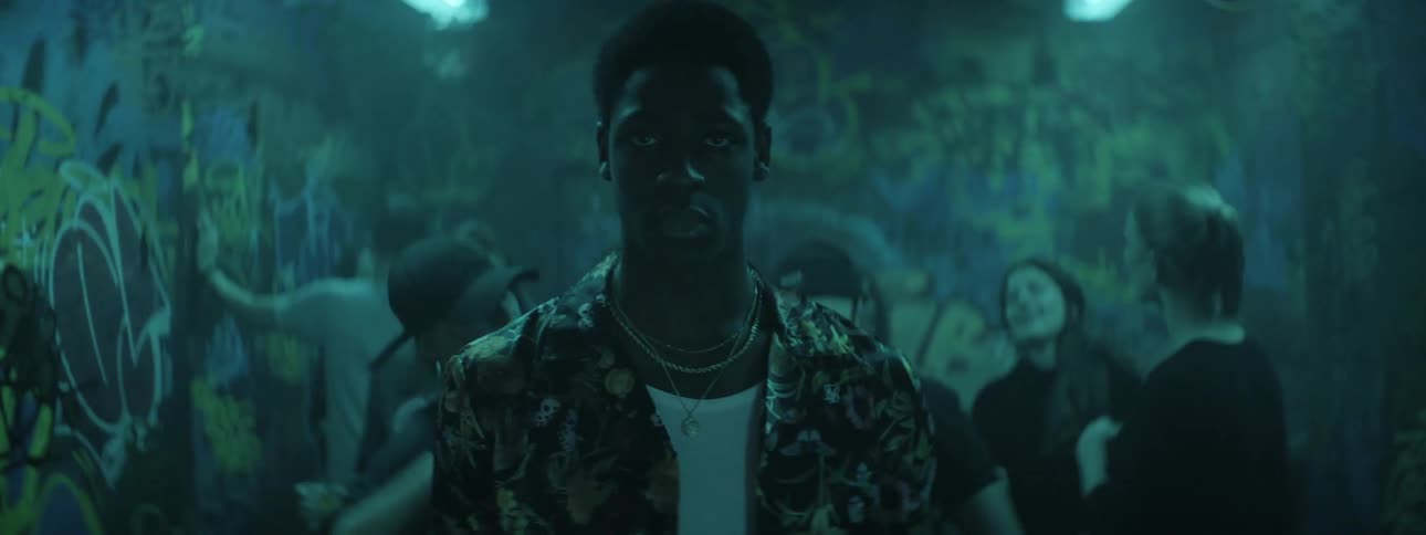 A male who is looking very serious walks through a group of people dancing in a room with flashing lights and graffiti covering the walls