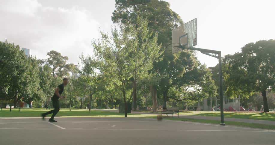A tall male dribbles a basketball and shoots a basket at an outdoor basketball court in a park