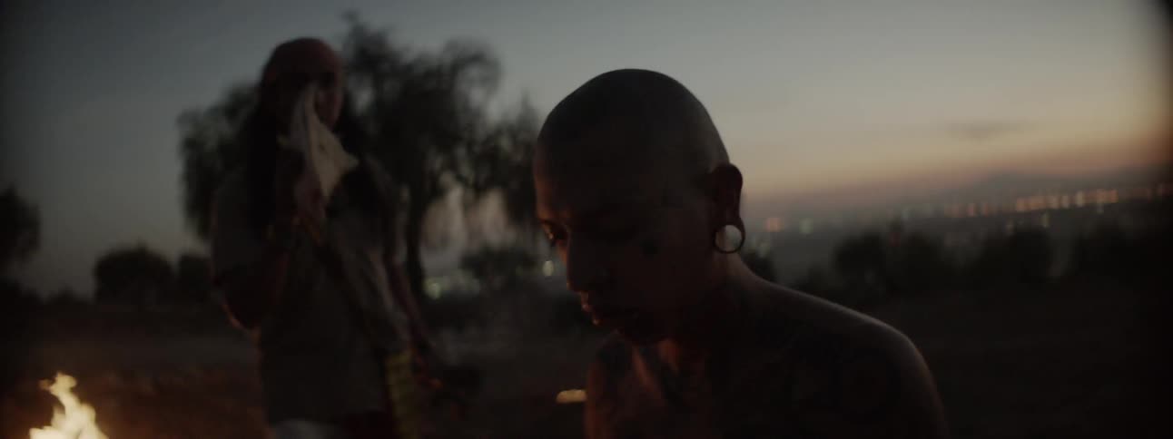 A man walks around a fire pit and takes a drink as another man sits there