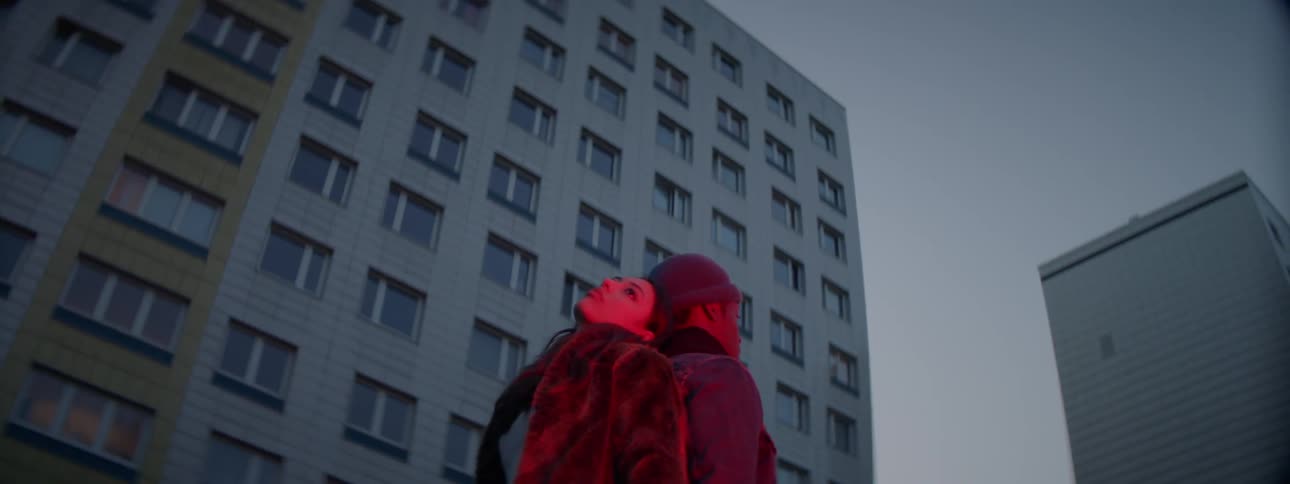 A man and woman stand with their backs against one another and look up at buildings