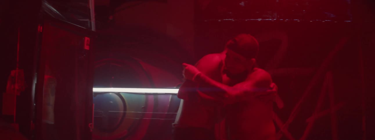 Two shirtless and tattooed men slap hands and then embrace in a darkened room