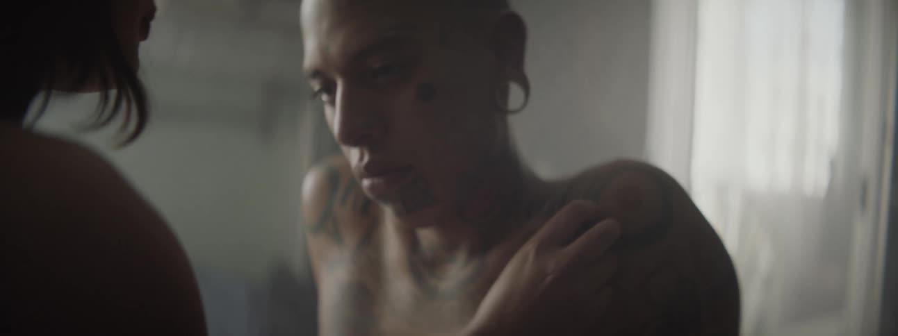A woman talks and prays as she touches the chest and shoulders of a man with many tattoos