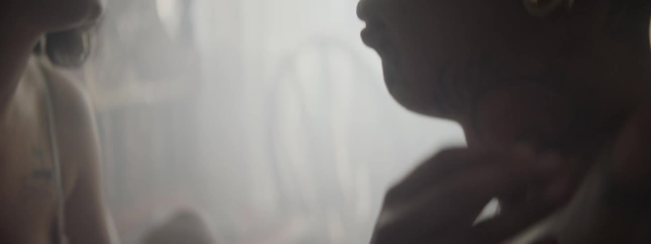A young woman touches an object to the face and chest of a heavily tattooed male