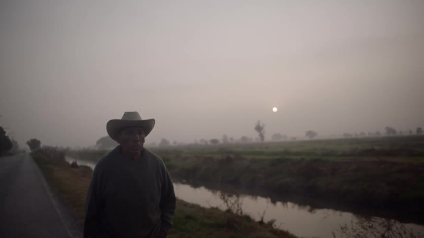 A man in a cowboy hat walks along a road that runs next to a canal