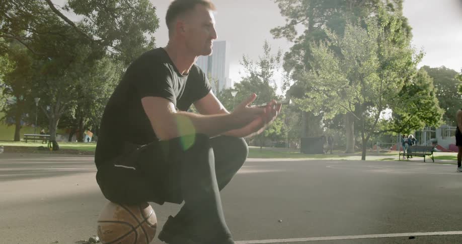 A young man sits on a basketball and makes a call on his cellphone