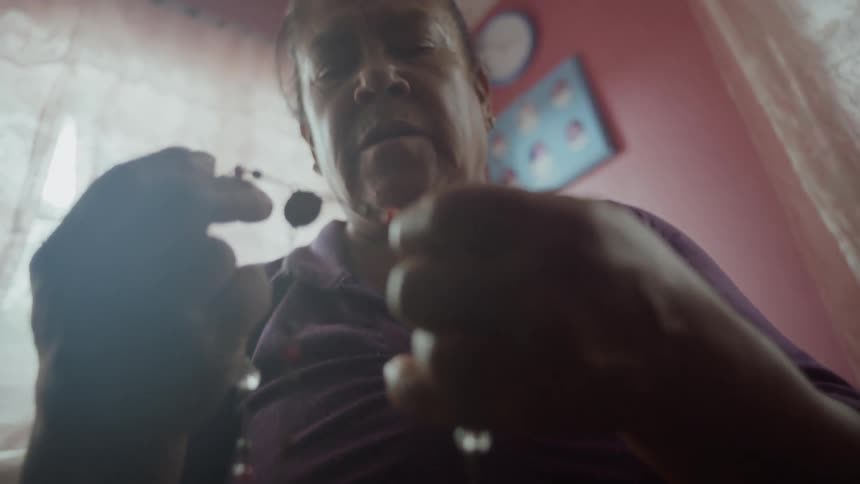 An elderly hispanic woman clutches a set of rosary beads as she sits and prays