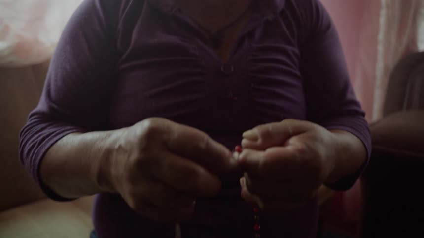An elderly hispanic woman prays while holding rosary beads in her hands