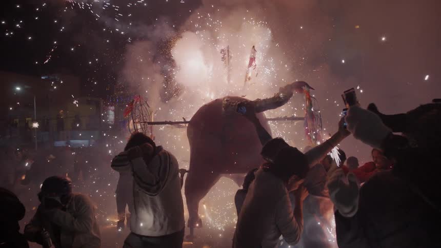 Crowd dances and takes pictures of fireworks display and bull shaped decoration