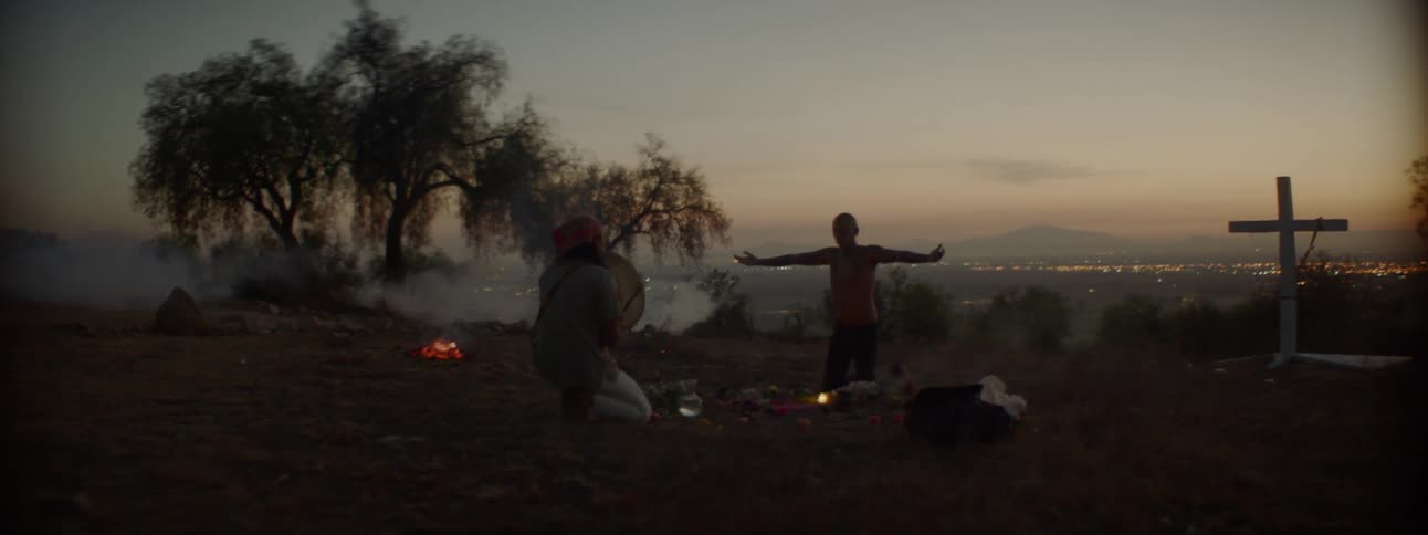 Two men sit by a fire while one holds his arms out and the other plays a drum