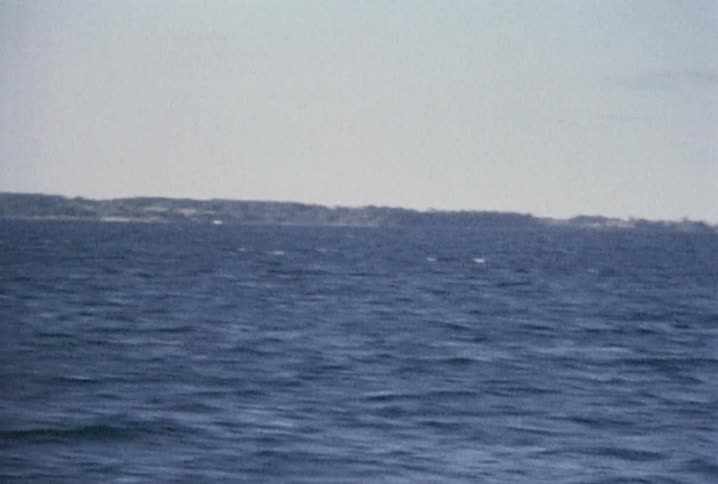Wide shot of choppy water, coastline, homes and a boat travelling in the distance