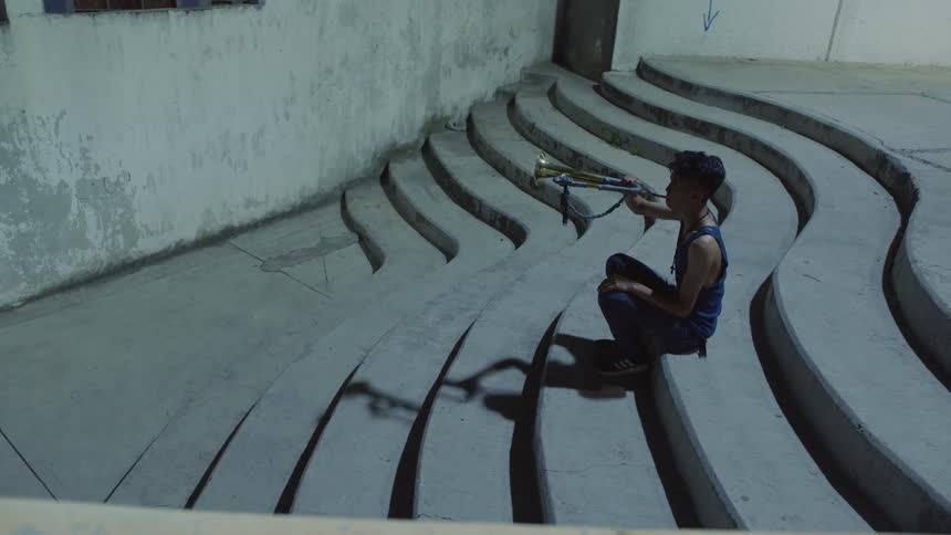 A teenage boy plays a trumpet by himself sitting on exterior steps
