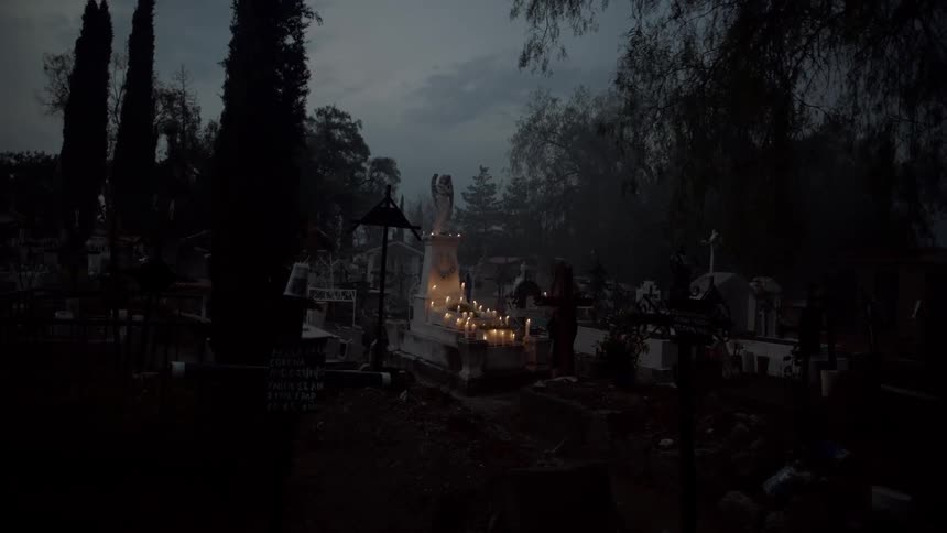 Woman kneels and leaves candle on a tomb in cemetery at dusk
