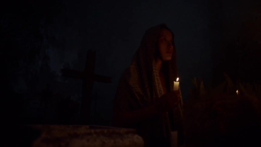 A young woman with a shawl over her head holds a candle in the darkness as she kneels beneath a tall cross
