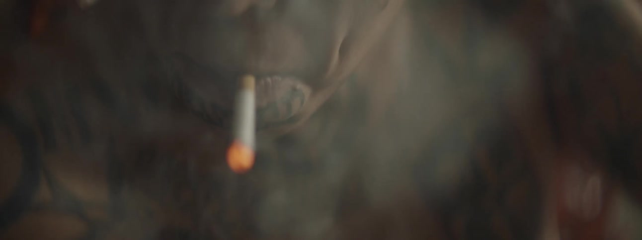 A hispanic mail with face tattoos and large earring lights and smokes a cigarette