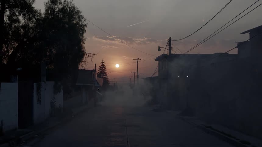 The sun hangs low in the sky above a dimly lit street with a cloud of smoke hovering in it