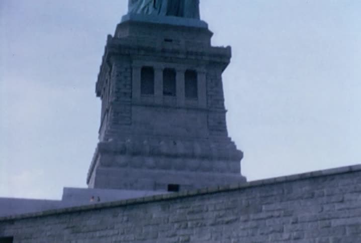 A young boy stands in the grass at the base of the statue of liberty and holds up a brochure