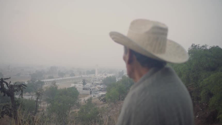 Man wearing cowboy hat stands on hill looking down on buildings