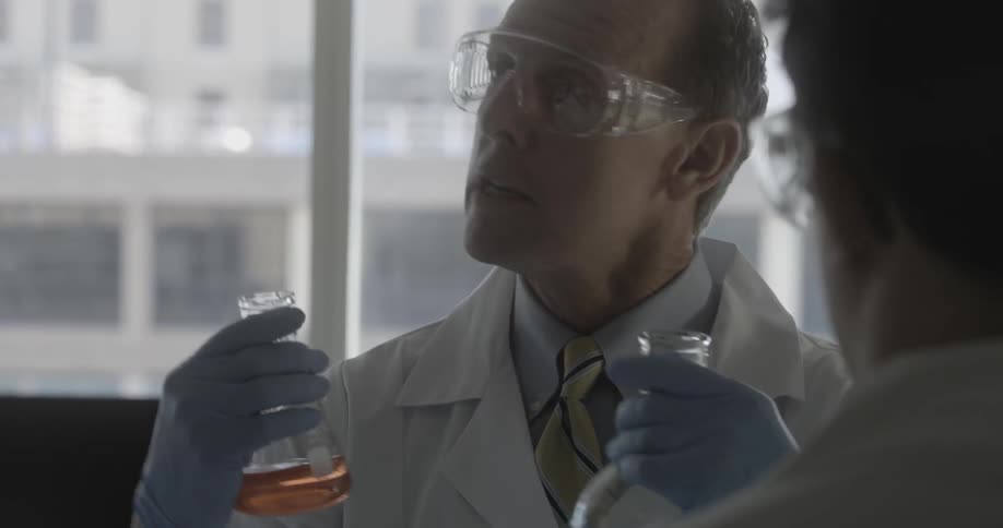 A scientist in a labcoat wearing safety goggles and gloves holds up two beakers as he speaks