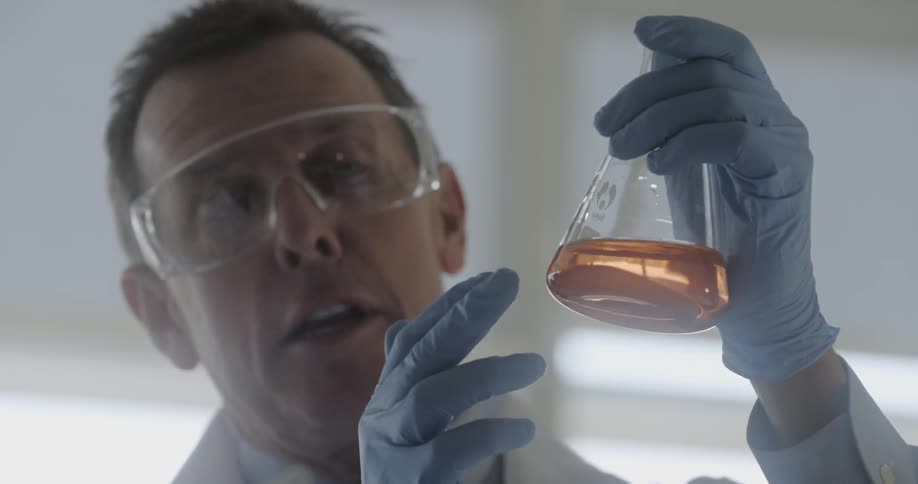 A scientist holds a beaker full of a clear orange fluid and talks to someone