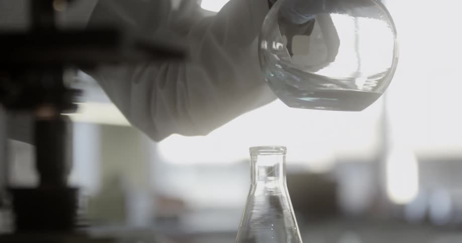 A chemist swirling a light blue liquid in a round bottom glass flask