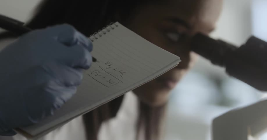 A woman looks through a microscope as a person wearing latex gloves takes notes on a pad