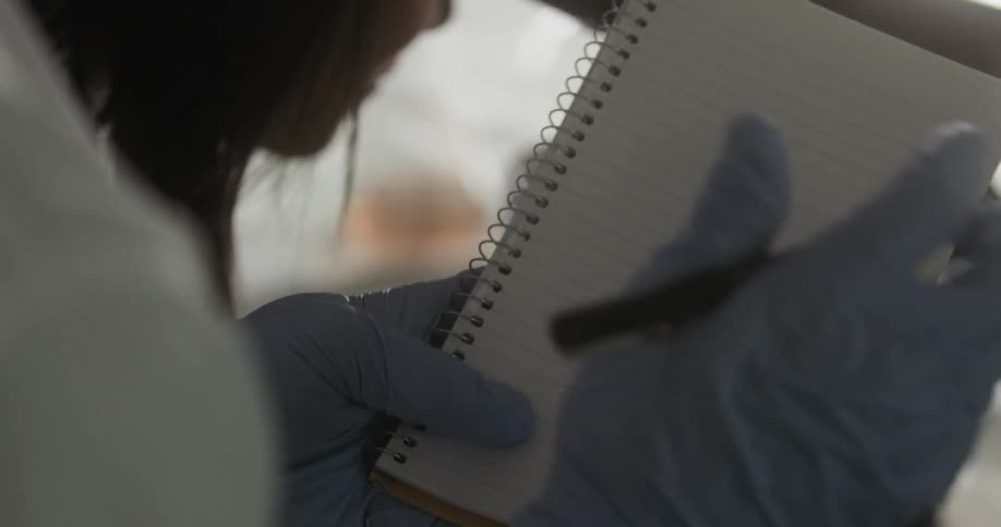 A person wearing latex gloves and a lab coat writes on a pad of paper