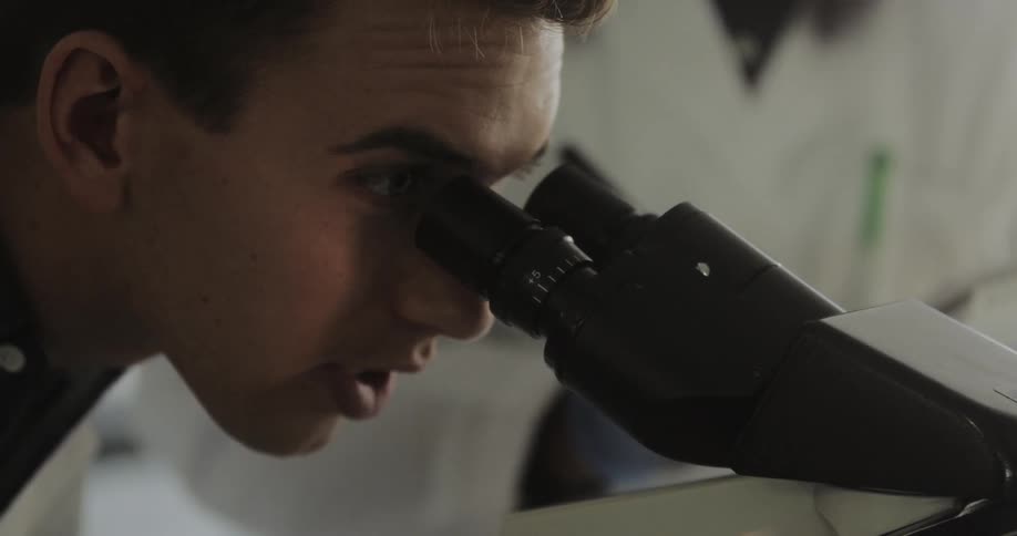 Scientist looks through microscope while talking to a colleague