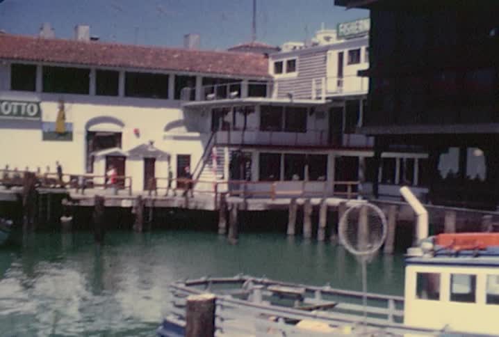 Old footage of boats docked in a harbor, a seaside restaurant and people milling around