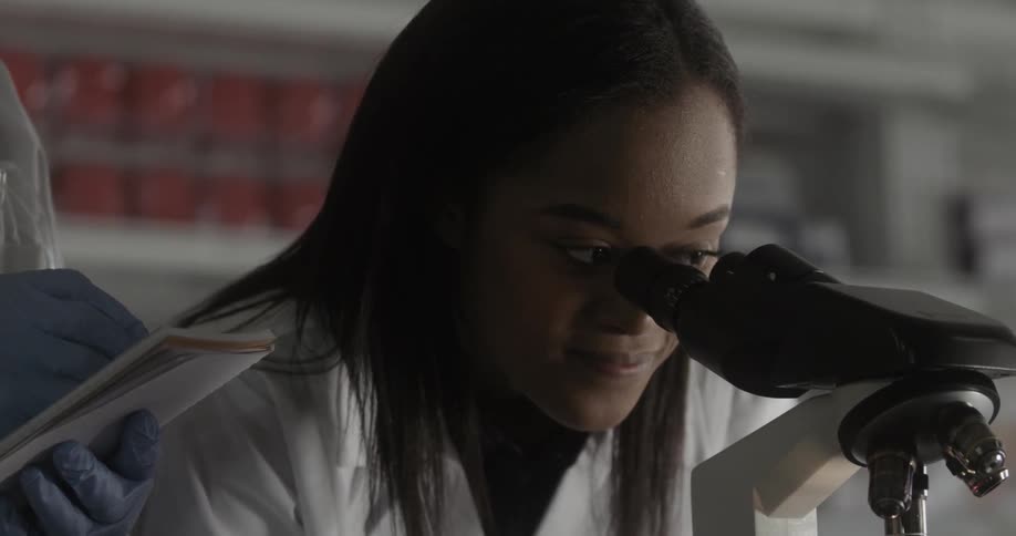 Scientist looks through microscope while colleague takes notes on laboratory