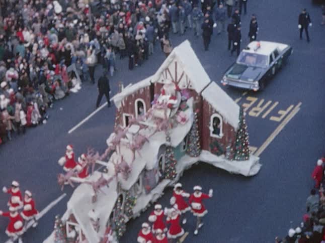 Women dressed in santa outfits walking alongside a gingerbread house float, with santa and his sleigh on top, during a parade