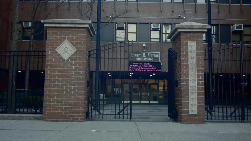 Half of the iron gate to a city high school building is open and there are lights on inside the building