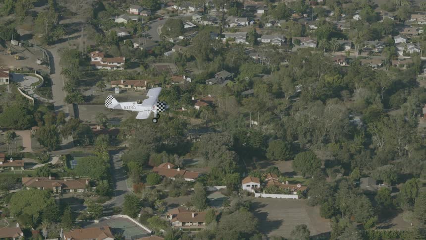 A bi-plane is flying over a suburban area during the day