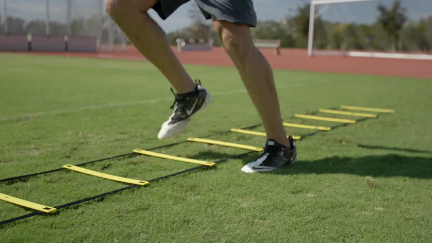A man jumping through a ladder thing that is laying on the grass of a sports field