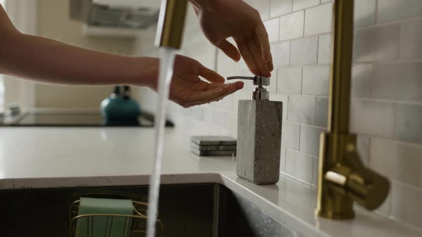 A woman washes her hands at the kitchen sink adding soap from a dispenser on the counter