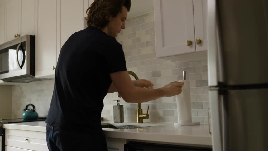 A man washes his hands and a kitchen sink and then dries them with a paper towel