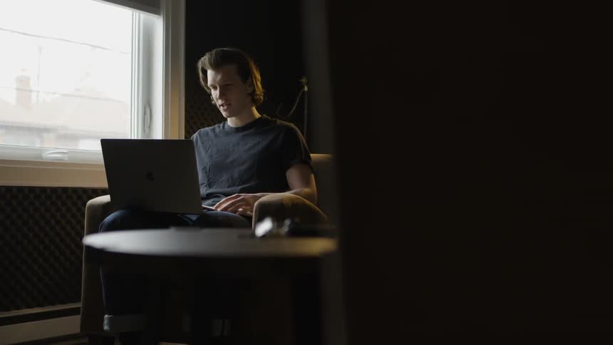 A man sits in a chair working on a laptop and touches his chin