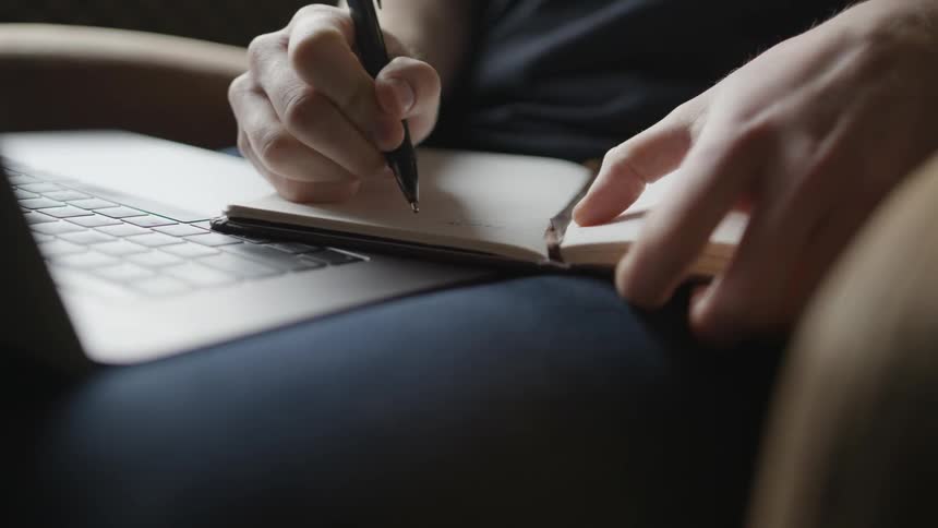 A person writing in a notebook with a laptop in his lap