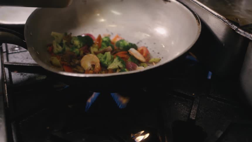 A frying pan on a stove with shrimp and vegetables while more shrimp is poured into the pan