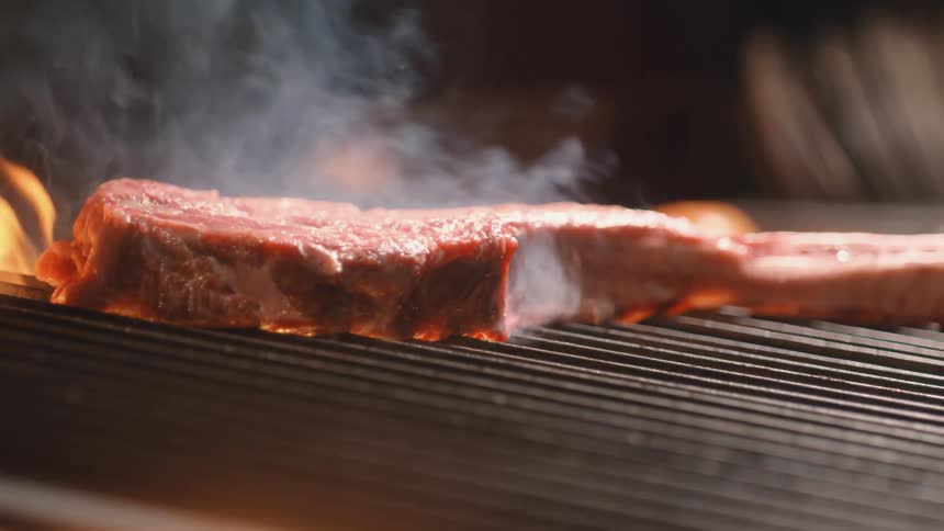 A piece of steak sits atop a grill as flames arise from underneath it and cook it