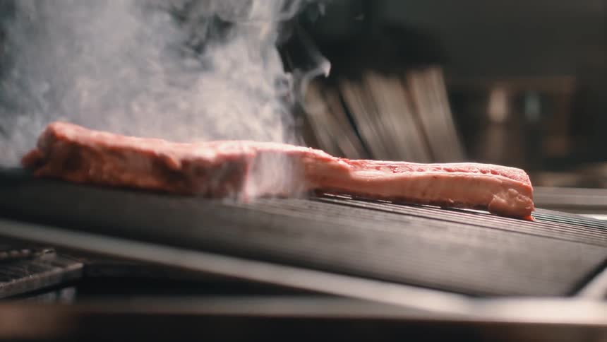 A person holds a large slab of steak and drops it onto a hot grill and it begins to steam and smoke as it cooks