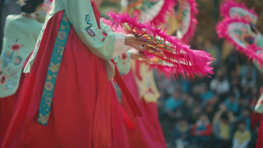 Female dancers in traditional costume and carrying fans perform outdoors in front of a crowd