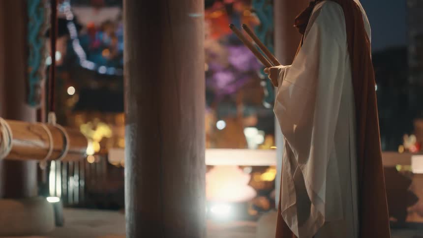 A robed man holding two sticks standing in shrine at night