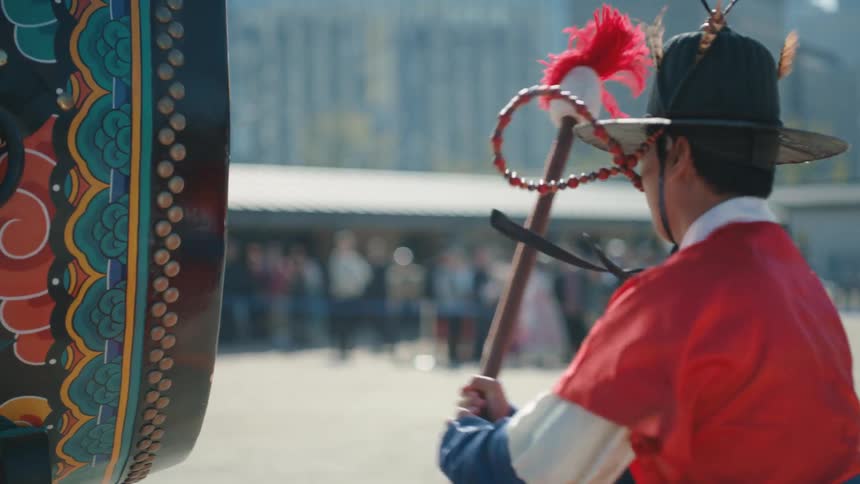 A man wearing a colorful uniform strikes a large drum with a mallet in front of a crowd of observers