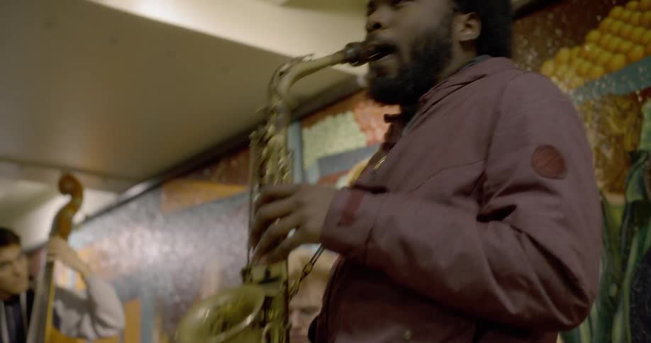 A black man plays a saxophone as other members of the band perform too