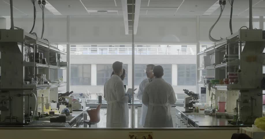Four scientists stand in a lab wearing white lab coats talking and taking notes