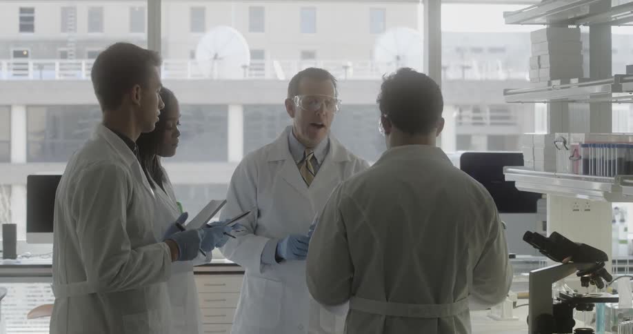 A group of scientists wearing white lab coats take notes while a male researcher speaks to them