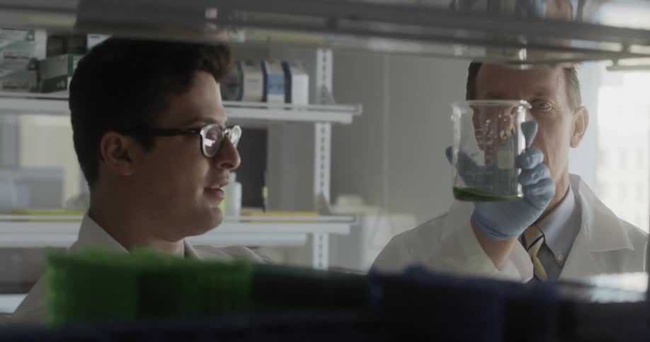 A man holds up a glass beaker with green liquid while talking with a colleague